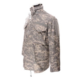 May include: A digital camouflage military jacket in shades of gray, tan, and green. The jacket features multiple pockets, a high collar with a zipper, and a drawstring at the bottom. The jacket is displayed on a mannequin.