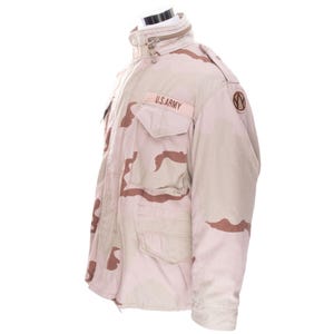 Könnte beinhalten: Eine militärische Tarnjacke in Beige und Braun mit dem Text "U.S.ARMY" auf der Brust. Die Jacke hat mehrere Taschen, einen Reißverschluss und einen hohen Kragen. Die Jacke wird auf einer Schaufensterpuppe präsentiert.