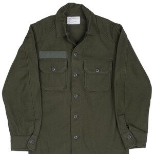May include: Olive green button-down shirt with two chest pockets and a small patch above the left pocket. The shirt has a collar and buttoned cuffs. A small tag is visible near the collar.