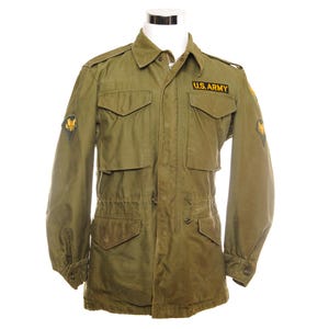 May include: An olive green military jacket with multiple pockets, a button-down front, and a drawstring waist. The jacket features a "U.S. ARMY" patch and two patches on the sleeves. The jacket is displayed on a white mannequin.