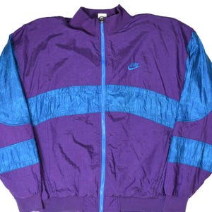 May include: A purple and blue windbreaker jacket featuring a full-length bright blue zipper, a high collar, and ribbed cuffs. The jacket displays a horizontal blue panel across the chest and sleeves, with the Nike logo embroidered in blue on the chest.