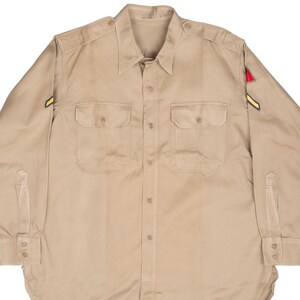 May include: A tan button-down shirt with two chest pockets and a collared neckline. The long-sleeved shirt has a military-style design, with patches on the sleeves. The shirt is made of a durable fabric and features button closures.