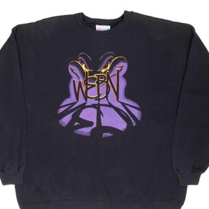 May include: Black sweatshirt featuring a purple and gold graphic design with the word "WEEN" in a stylized font. The graphic has a surreal, eye-like shape. The sweatshirt is a crew neck style.