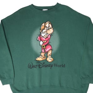 Könnte beinhalten: Ein dunkelgrünes Sweatshirt mit Rundhalsausschnitt und einer Grafik der Figur Grumpy aus Schneewittchen mit verschränkten Armen. Unter der Figur ist der Text Walt Disney World in Schwarz gedruckt. Das Sweatshirt hat lange Ärmel und einen Rippkragen.