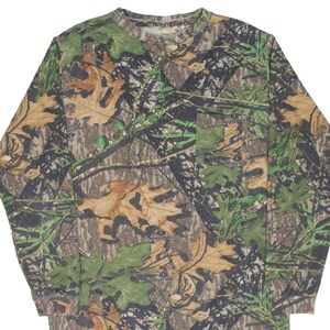Puede incluir: Camiseta de camuflaje de manga larga con cuello redondo y bolsillo en el pecho izquierdo. La camiseta presenta un estampado de hojas y ramas marrones, verdes y beiges. La marca "Mossy Oak" es visible en el bolsillo.