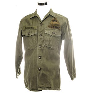 Puede incluir: Camisa militar verde oliva con mangas largas, dos bolsillos en el pecho y cierres de botones. Cuenta con un parche que dice "HELL ON WHEELS U.S. ARMY" y una etiqueta con el nombre "ESPENSHADE".
