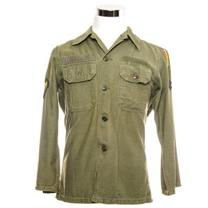 May include: Olive green military shirt with long sleeves, two chest pockets, and button closures. The name "BUFFORD" is displayed above the left chest pocket. Patches are visible on the sleeves. The shirt is displayed on a white mannequin.