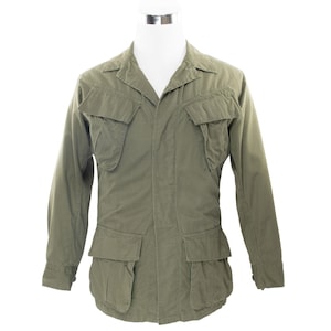 Op de afbeelding: Olijfgroene jas in militaire stijl met vier voorzakken en een kraag. De jas met lange mouwen wordt gepresenteerd op een witte etalagepop. De jas heeft een knoopsluiting en een getailleerde pasvorm.