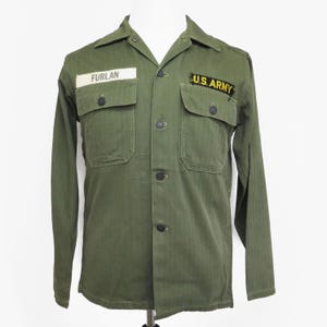 May include: Olive green military-style shirt with two chest pockets and black buttons. The shirt features a name tag that reads "FURLAN" and a patch that reads "U.S. ARMY". The long-sleeved shirt has a collared neckline.