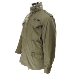 Puede incluir: Chaqueta militar verde oliva con múltiples bolsillos y cuello alto. La chaqueta presenta un parche de "U.S. ARMY" en el pecho. La chaqueta se exhibe en un maniquí.