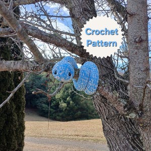 Puede incluir: Dos peluches de ganchillo azules y blancos con forma de serpiente cuelgan de una rama de árbol. También se ve un letrero blanco con las palabras "Crochet Pattern". El fondo muestra un campo y árboles.
