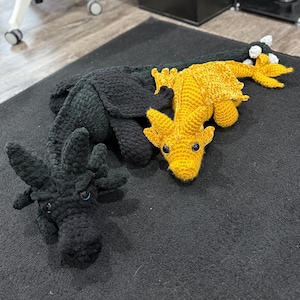 May include: Two crocheted dragon toys. One is black, the other is yellow. The black dragon has blue eyes and a spiky head. The yellow dragon has a long tail and small horns. Both are lying on a black surface.