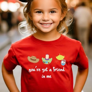 May include: Red short-sleeved t-shirt with a cartoon image of Woody, Buzz Lightyear, and Alien from Toy Story. The shirt has the text "you've got a friend in me" printed on it.
