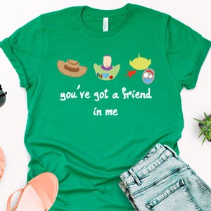 May include: Green t-shirt with a white graphic of characters from the Toy Story movie. The graphic includes Woody, Buzz Lightyear, and the alien. The text on the shirt reads "you've got a friend in me."