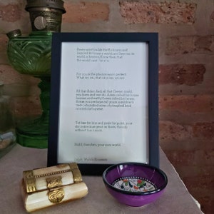 Pode incluir: Uma impress&atilde;o emoldurada com uma moldura preta exibindo uma cita&ccedil;&atilde;o de Ralph Waldo Emerson. Uma pequena caixa decorativa com detalhes dourados e uma tigela roxa com um design floral est&atilde;o em frente &agrave; moldura. Uma l&acirc;mpada de vidro verde &eacute; vis&iacute;vel.