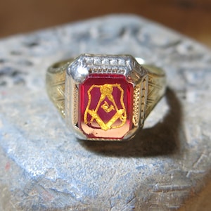 May include: A gold-toned ring with a rectangular, ruby-red center stone. The stone features a gold emblem. The ring has a detailed design around the stone and on the band. The ring is set on a gray, textured stone.