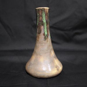 May include: A ceramic vase with a narrow neck and a wide, rounded base. The surface features a textured, mottled finish in shades of tan, beige, and brown, with a distinct green glaze dripping vertically from the rim down the neck of the vessel.