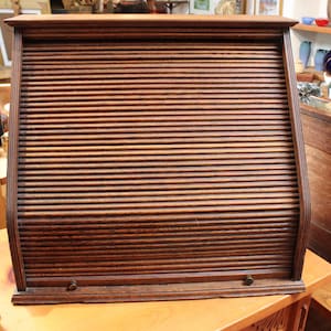 May include: A dark brown wooden roll-top desk organizer featuring a slatted tambour door that covers the front. The unit has a flat top surface and two small circular wooden knobs at the bottom edge for lifting the sliding door mechanism.