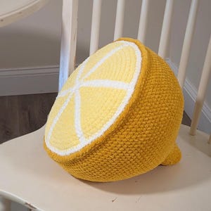 May include: A crocheted lemon-shaped cushion in shades of yellow, white, and a darker yellow-orange. The cushion is a half-lemon slice design, with white segments and a textured surface. It sits on a white chair.