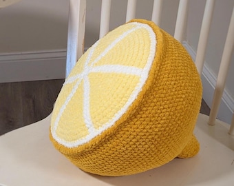 May include: A crocheted lemon-shaped pillow in shades of yellow, white, and a darker yellow-orange. The pillow is a half-lemon slice design, with white segments and a textured surface. It sits on a white chair.