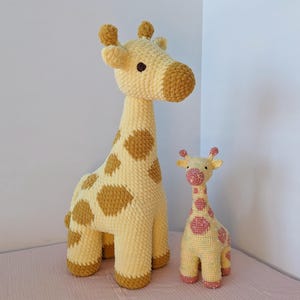 May include: Two handmade crochet giraffe plush toys. The larger giraffe is yellow with brown spots and a tan snout. The smaller giraffe is yellow with pink spots and a pink snout. Both have small, dark eyes and are standing upright.