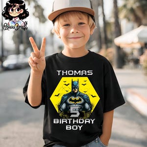 May include: Black t-shirt featuring a Batman graphic with the name "Thomas" and the text "5 BIRTHDAY BOY". The design includes a yellow hexagon with a Batman illustration and bat silhouettes. The boy is wearing a baseball cap.