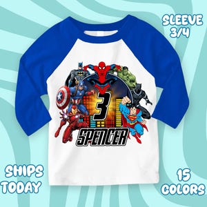 May include: A white and blue raglan style shirt featuring a group of superheroes. The shirt has 3/4 sleeves and the number "3" with the name "Spencer" printed on the front. The shirt also has the text "15 COLORS" and "SHIPS TODAY".