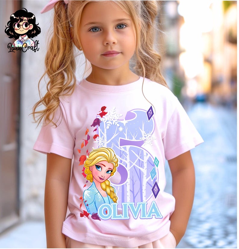 Puede incluir: Una camiseta rosa claro con un gr&aacute;fico de Elsa de Frozen, el n&uacute;mero 5 y el nombre "Olivia" en azul claro. El dise&ntilde;o incluye copos de nieve y una escena de bosque. La camiseta la lleva una ni&ntilde;a.