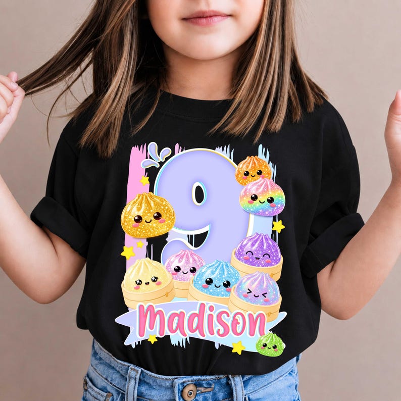 May include: A black t-shirt featuring a large light purple number nine graphic surrounded by colorful, smiling cartoon dumplings in shades of yellow, orange, pink, blue, and purple. The name Madison is printed in pink script at the bottom.