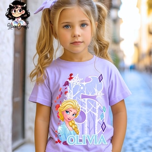 Puede incluir: Una camiseta lavanda con un gr&aacute;fico de Elsa de Frozen, el n&uacute;mero 5 y el nombre Olivia en azul. El dise&ntilde;o incluye copos de nieve y elementos forestales. La ni&ntilde;a lleva un lazo lavanda a juego.