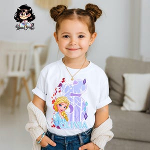 Puede incluir: Una ni&ntilde;a peque&ntilde;a lleva una camiseta blanca con un gr&aacute;fico de Elsa de Frozen, el n&uacute;mero 5 y el nombre "Olivia" en azul. El dise&ntilde;o incluye detalles en morado y rojo. Lleva un collar dorado y una pulsera.