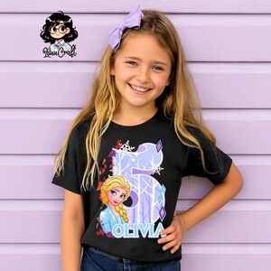 Puede incluir: Camiseta negra con un gr&aacute;fico de Elsa de Frozen, el n&uacute;mero 5 y el nombre Olivia en blanco. El dise&ntilde;o incluye copos de nieve y una escena de bosque invernal. La ni&ntilde;a lleva un lazo morado.
