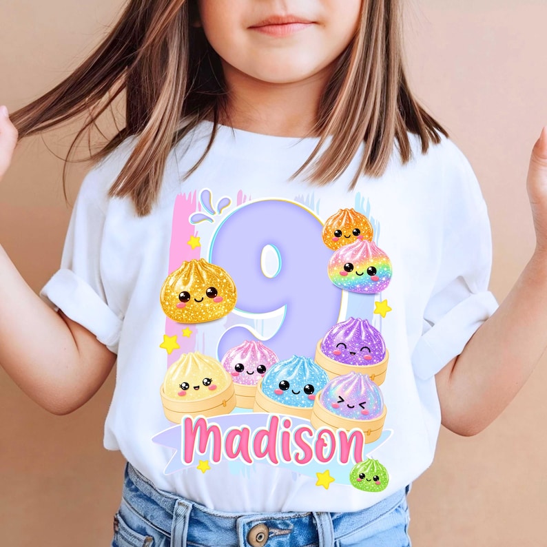 May include: A white t-shirt featuring a large light purple number nine graphic surrounded by colorful, smiling, glittery dumpling illustrations in bamboo steamers. The name Madison is printed in pink script below the number, accented by small yellow stars.