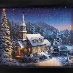 Może przedstawiać: Oprawione puzzle przedstawiające zimową scenę z kościołem i choinką. Kościół z wysoką wieżą jest oświetlony od wewnątrz, a po lewej stronie stoi udekorowana choinka. Puzzle osadzone są na tle gwiaździstego nieba i ośnieżonych drzew.