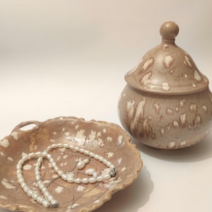 Op de afbeelding: Een bruine keramische schaal met een parelketting erop. De schaal heeft een gegolfde rand en een handvat aan elke kant. De schaal staat op een wit oppervlak.