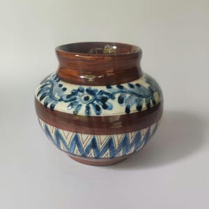 Op de afbeelding: Een kleine, ronde keramische vaas met een bruine glazuur en blauwe en witte bloemenontwerpen. De vaas heeft een geometrisch patroon rond de bodem.