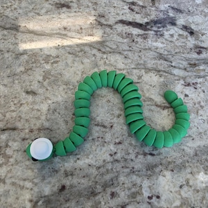 May include: A green articulated toy snake with a segmented body and a white circular eye detail on its head. The toy features a flexible, repeating structure that allows it to bend and curve into various positions while maintaining its shape.