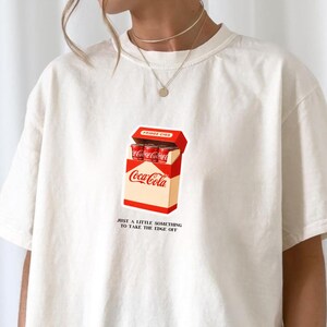 以下が含まれることがあります： クリーム色のTシャツ。赤い缶が入ったコカ・コーラの箱のグラフィックが特徴です。箱は開いており、中の缶が見えます。箱の下には「JUST A LITTLE SOMETHING TO TAKE THE EDGE OFF」と書かれています。