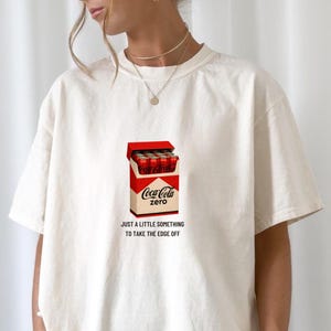 Op de afbeelding: Crèmekleurig T-shirt met een afbeelding van een Coca-Cola Zero-doos in rood en wit. De tekst "JUST A LITTLE SOMETHING TO TAKE THE EDGE OFF" staat eronder. Het shirt heeft een losse pasvorm.