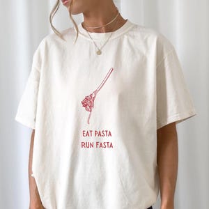 Peut inclure: T-shirt crème avec un dessin rouge de pâtes sur une fourchette et le texte "EAT PASTA RUN FASTA". Le t-shirt est ample et à manches courtes. Le modèle porte un short noir.