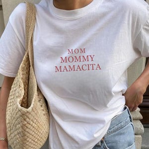 Pu&ograve; includere: T-shirt bianco con le parole "MOM MOMMY MAMACITA" in lettere rosse. La maglietta &egrave; abbinata a jeans azzurri e una borsa tote beige intrecciata. Una tazza di caff&egrave; nera &egrave; tenuta in mano.