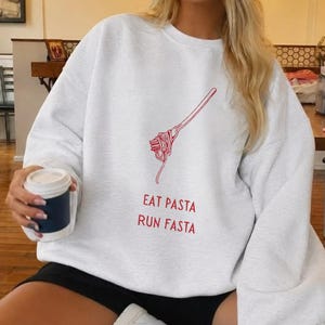 May include: A light grey crewneck sweatshirt featuring a red line-art graphic of a fork twirling spaghetti. Below the graphic, the text reads EAT PASTA RUN FASTA in red capital letters. The sweatshirt has long sleeves and a relaxed fit.