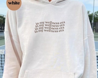 May include: An off-white hooded sweatshirt featuring a front kangaroo pocket and the text "in my wellness era" printed in brown ink four times in a stacked, slightly wavy alignment across the centre of the chest area.