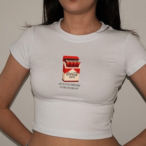May include: White crop top featuring a graphic of a Coca-Cola Zero pack. The pack is red and white with the Coca-Cola Zero logo and the text "JUST A LITTLE SOMETHING TO TAKE THE EDGE OFF".