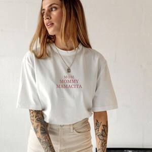 Pu&ograve; includere: T-shirt bianco con le parole "MOM MOMMY MAMACITA" in rosso. La maglietta &egrave; in stile casual con maniche corte. La modella indossa una collana d'argento e pantaloni colorati chiari.