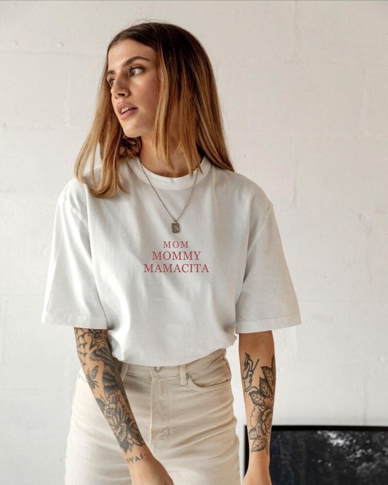 Pu&ograve; includere: T-shirt bianco con le parole "MOM MOMMY MAMACITA" in rosso. La maglietta &egrave; in stile casual con maniche corte. La modella indossa una collana d'argento e pantaloni colorati chiari.