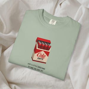 May include: A sage green crewneck t-shirt featuring a graphic of a red carton filled with Coke Zero cans. Below the carton, the text reads: JUST A LITTLE SOMETHING TO TAKE THE EDGE OFF. The shirt has a Comfort Colors label at the inner neckline.