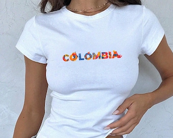 May include: White t-shirt with the word "COLOMBIA" in colorful, floral-style lettering. The shirt has short sleeves and a fitted silhouette. The design is centered on the chest.