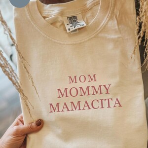 Pu&ograve; includere: T-shirt Comfort Colors color sabbia con le parole "MOM MOMMY MAMACITA" stampate in marrone rossiccio. La maglietta &egrave; piegata e tenuta in mano. L'etichetta recita "S-P-CH".