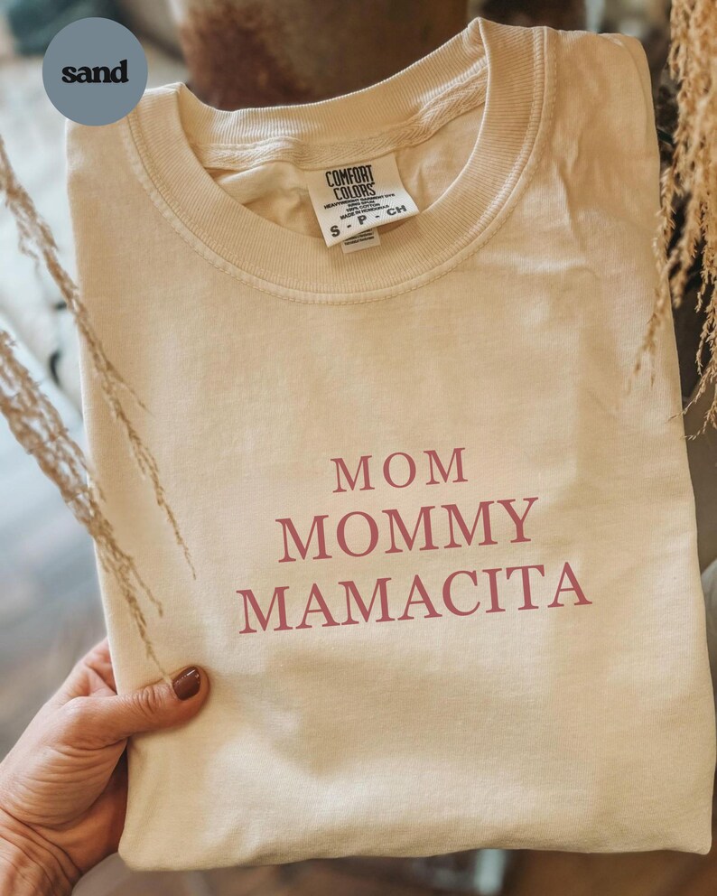Pu&ograve; includere: T-shirt Comfort Colors color sabbia con le parole "MOM MOMMY MAMACITA" stampate in marrone rossiccio. La maglietta &egrave; piegata e tenuta in mano. L'etichetta recita "S-P-CH".